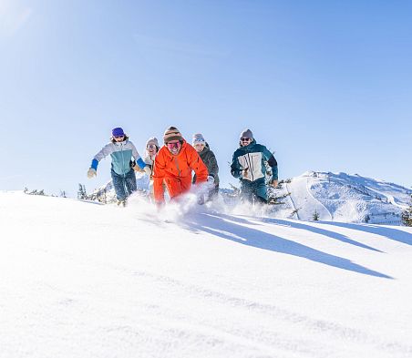Fünf Personen laufen durch Tiefschnee. Im Hintergrund sieht man klein eine Skipiste. Eine der Personen in einem orangen Skianzug ist am weitesten vorne.