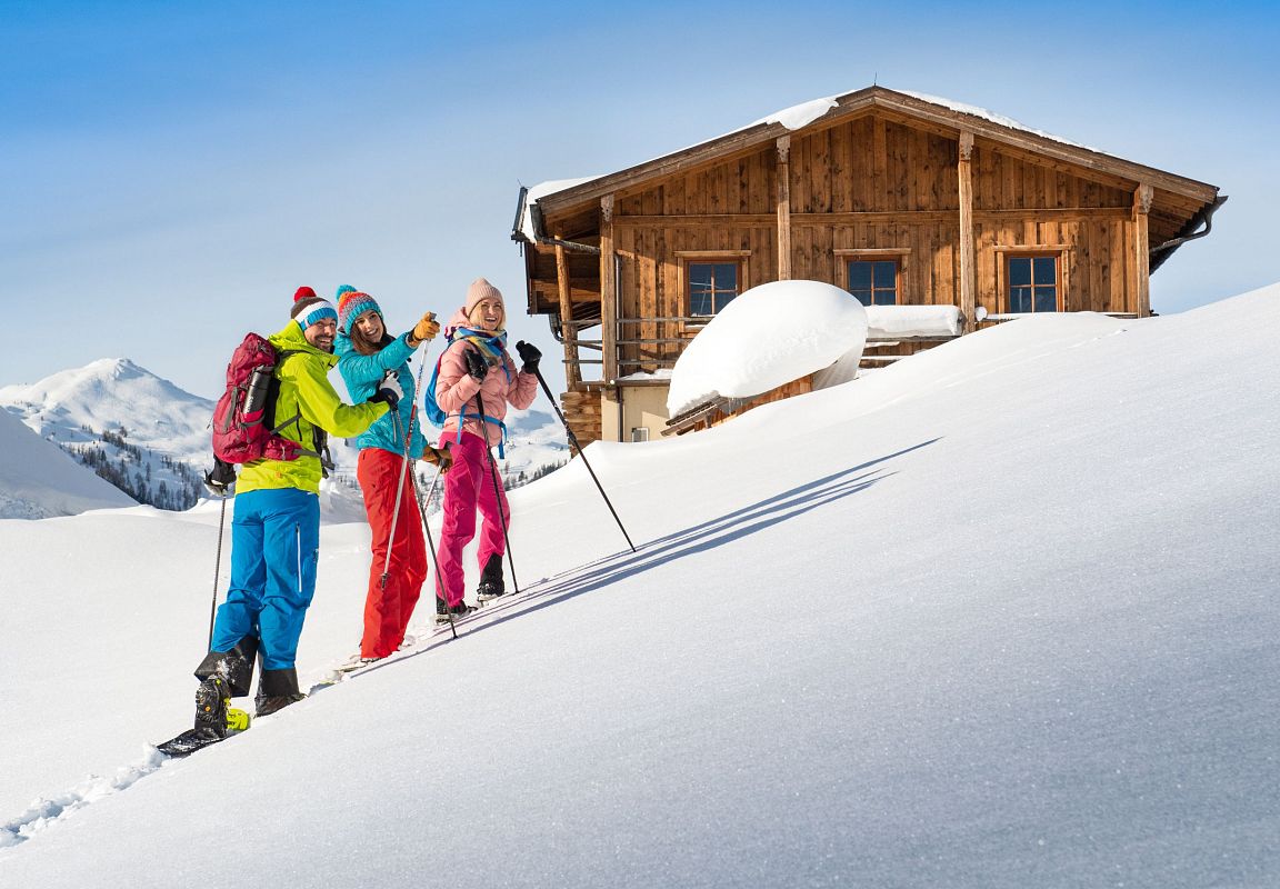 Drei Schneeschuhwanderer  nähern sich einer verschneiten Berghütte – Naturerlebnis in Flachau, Salzburger Land.