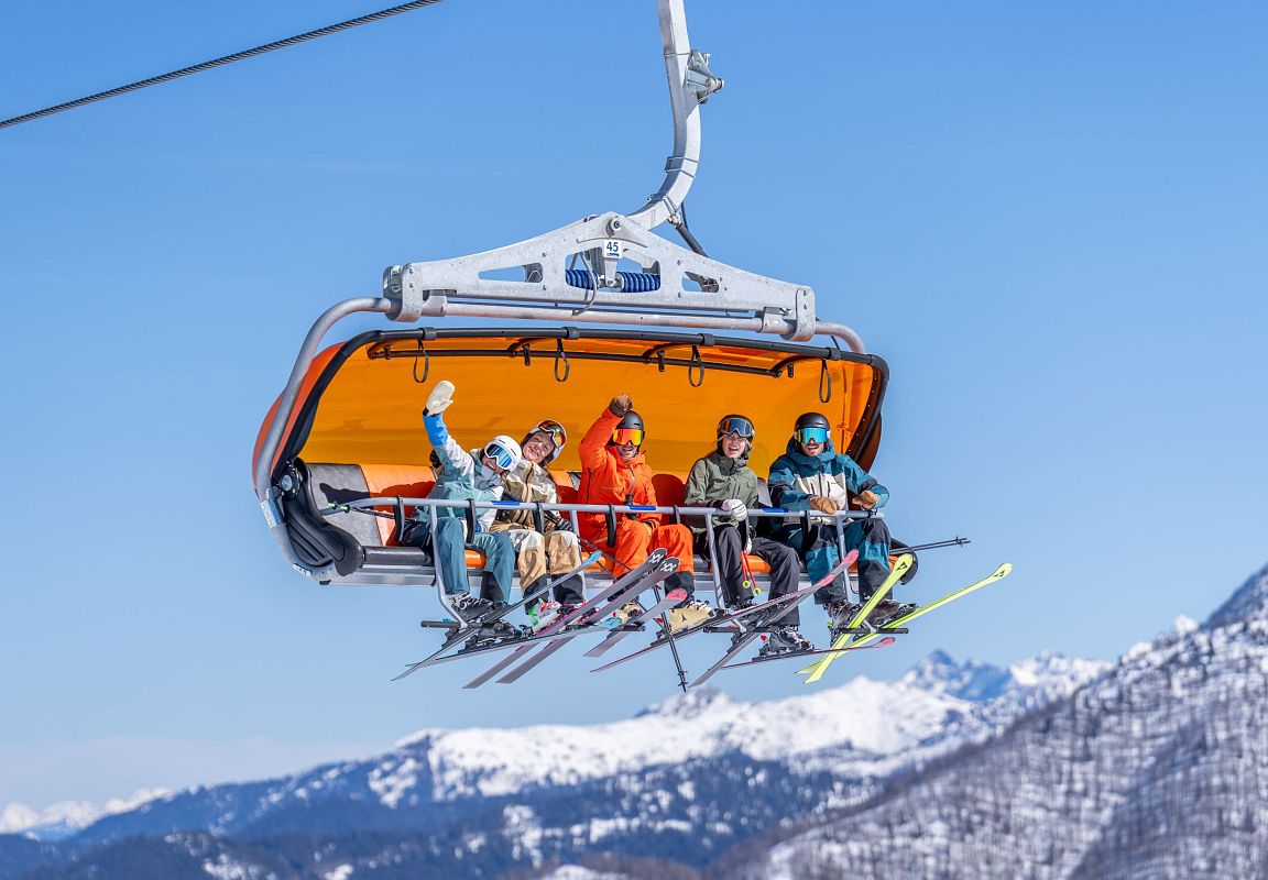 Gruppe von Skifahrern fährt im Sessellift mit orangener Wetterschutzhaube, strahlende Gesichter bei blauem Himmel und verschneiten Bergen. Skigenuss in Flachau.