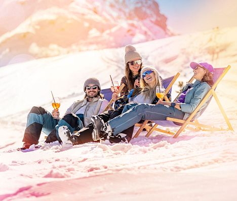Vier Personen sitzen auf Liegestühlen im Schnee mit einem Getränk in der Hand. Im Hintergrund sieht man eine Berggipfel. Das Bild hat einen bunten Filter.