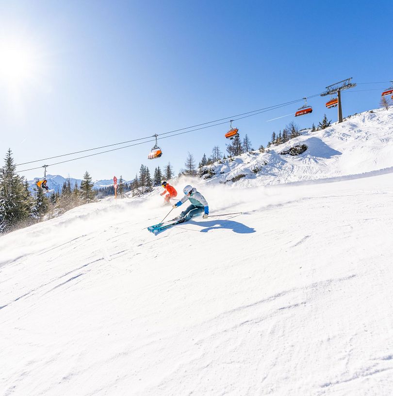 Zwei Skifahrer bei der Abfahrt vor einem orangen Sessellift. Beste Pistenverhältnisse und blauer Himmel.