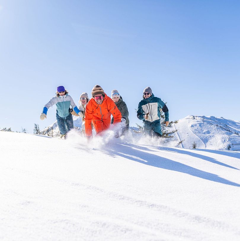 Fünf Personen laufen durch Tiefschnee. Im Hintergrund sieht man klein eine Skipiste. Eine der Personen in einem orangen Skianzug ist am weitesten vorne.