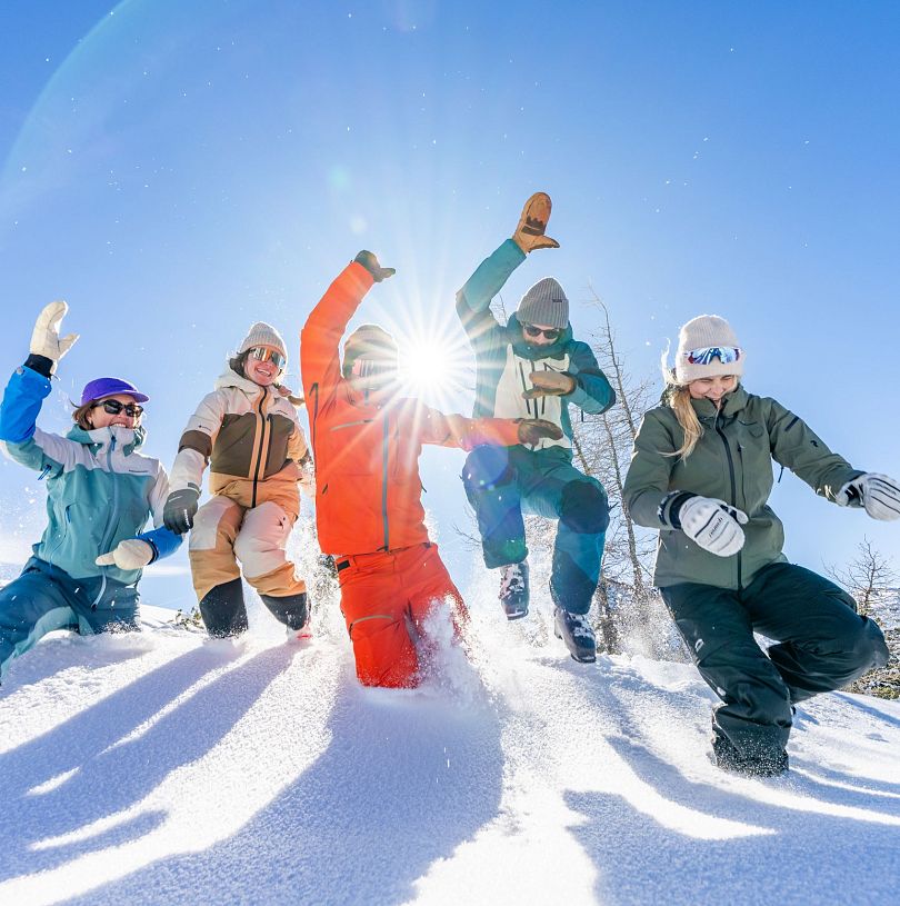 Fünf Personen springen in tiefen Schnee in Flachau am Berg. Die Sonne leichtet von hinten zwischen zwei der Personen durch.