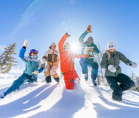Fünf Personen springen in tiefen Schnee in Flachau am Berg. Die Sonne leichtet von hinten zwischen zwei der Personen durch.