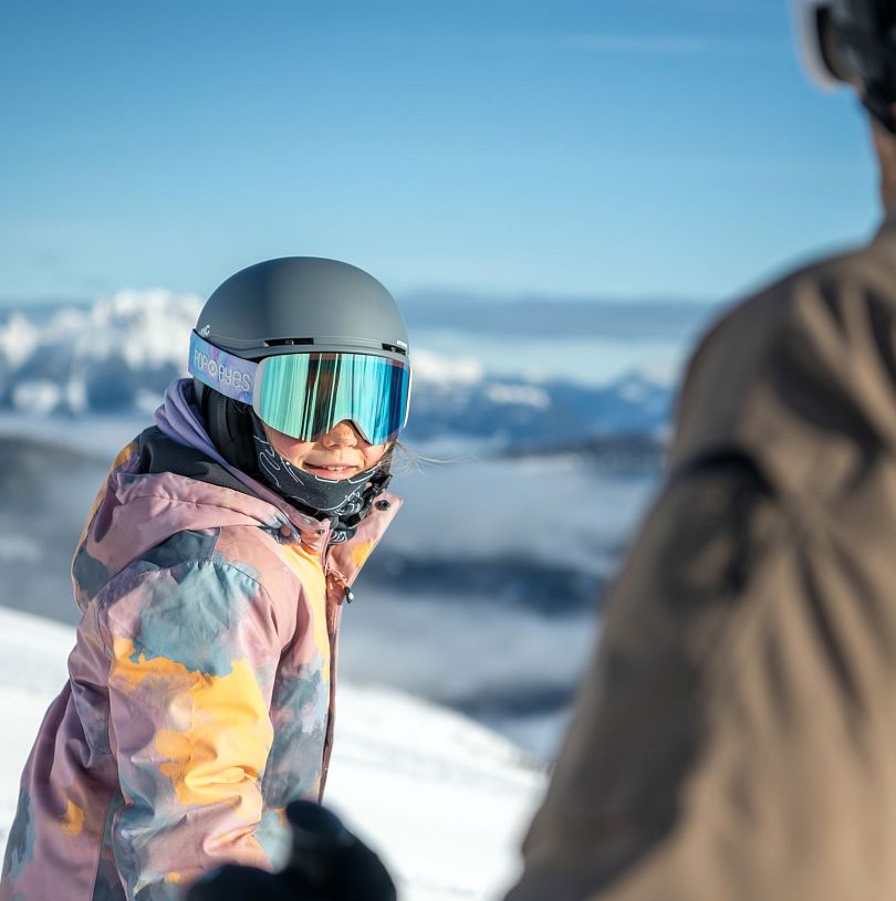 Kind mit Helm und Skibrille auf der Skipiste vor verschneitem Alpenpanorama in Flachau.