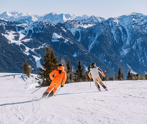 Zwei Skifahrer fahren eine Abfahrt hinunter. Man sieht eine wunderschöne Bergkulisse.