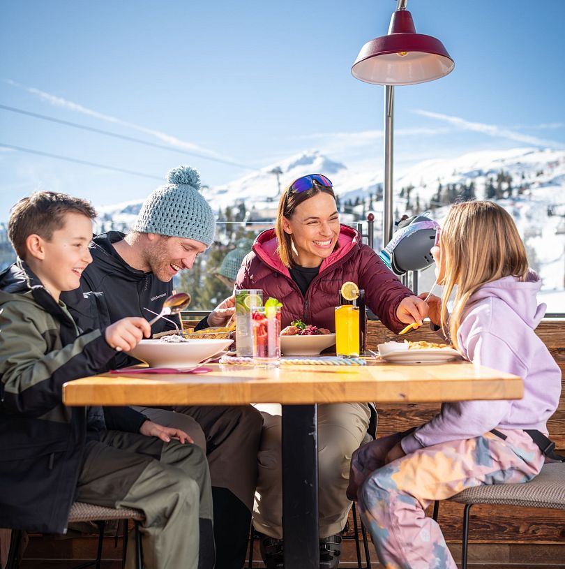 Familie beim gemeinsamen Essen auf einer Sonnenterrasse mit Blick auf die verschneiten Berge in Flachau.