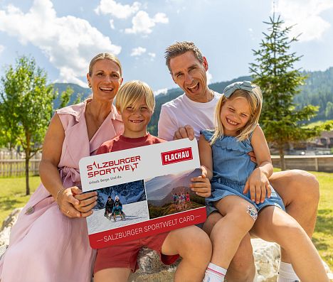 Eine Familie mit zwei Kindern hält eine grosse Version der Flachau Sommer Card in der Hand und lacht in die Kamera.