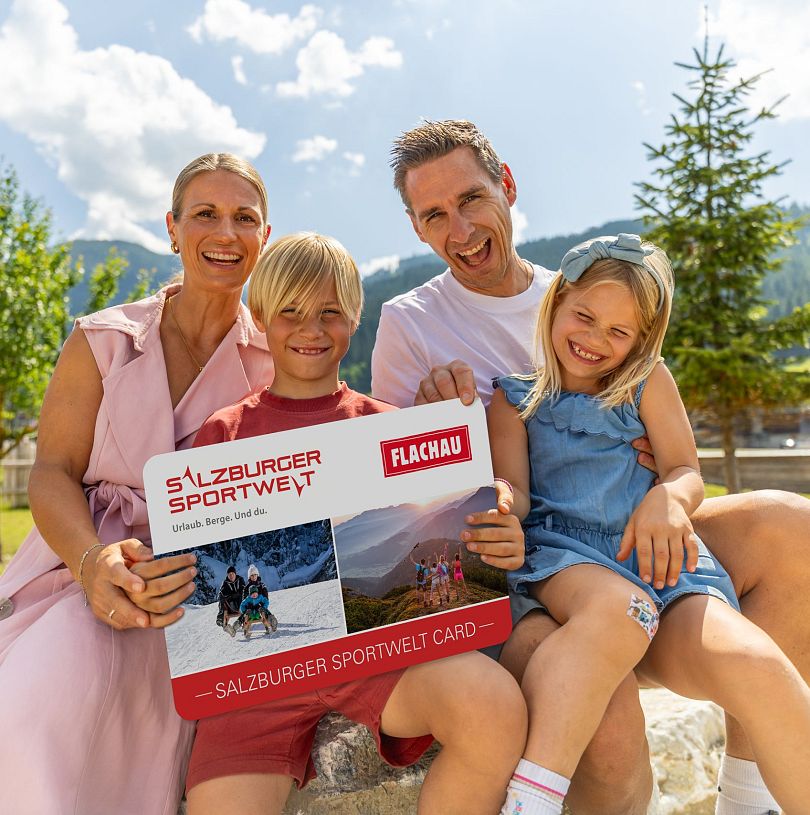 Eine Familie mit zwei Kindern hält eine grosse Version der Flachau Sommer Card in der Hand und lacht in die Kamera.
