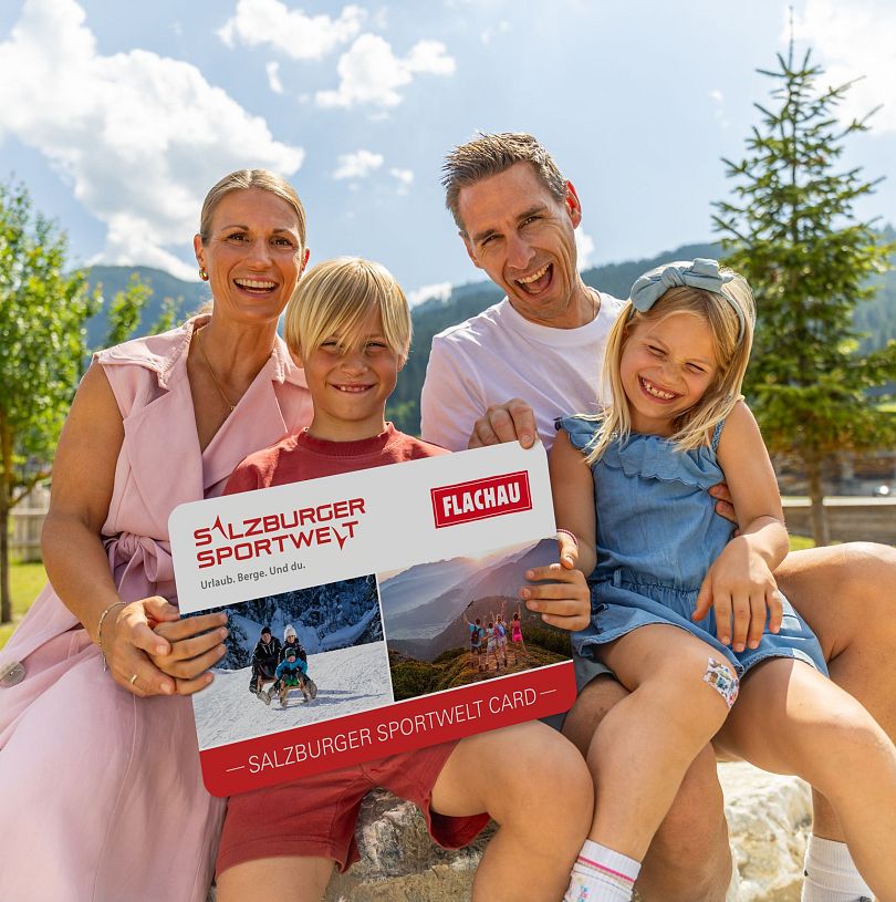 Eine Familie mit zwei Kindern hält eine grosse Version der Flachau Sommer Card in der Hand und lacht in die Kamera.