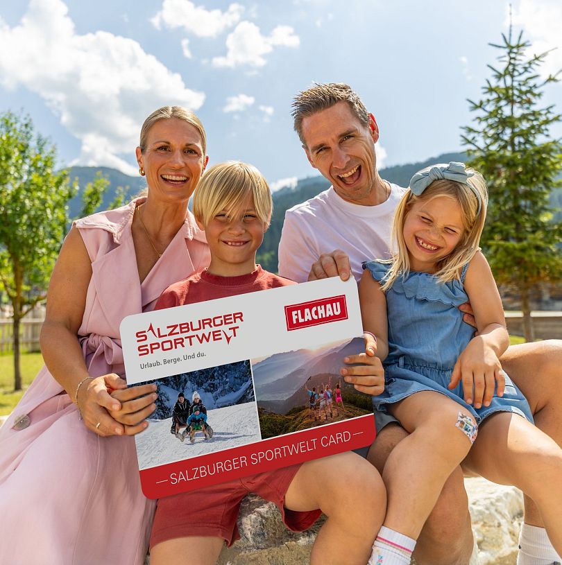 Eine Familie mit zwei Kindern hält eine grosse Version der Flachau Sommer Card in der Hand und lacht in die Kamera.