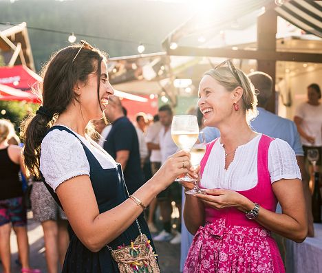 Zwei Frauen in Dirndl stoßen bei sommerlichem Gassenfest in Flachau, Salzburger Land, mit Wein an.