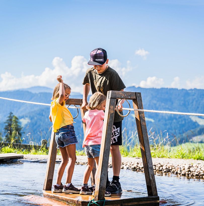 Kinder überqueren auf dem Floß den kleinen See - eine der beliebtesten Spielstationen auf Flori's Erlebnispfad in Flachau.