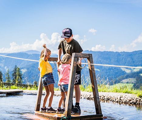 Kinder überqueren auf dem Floß den kleinen See - eine der beliebtesten Spielstationen auf Flori's Erlebnispfad in Flachau.