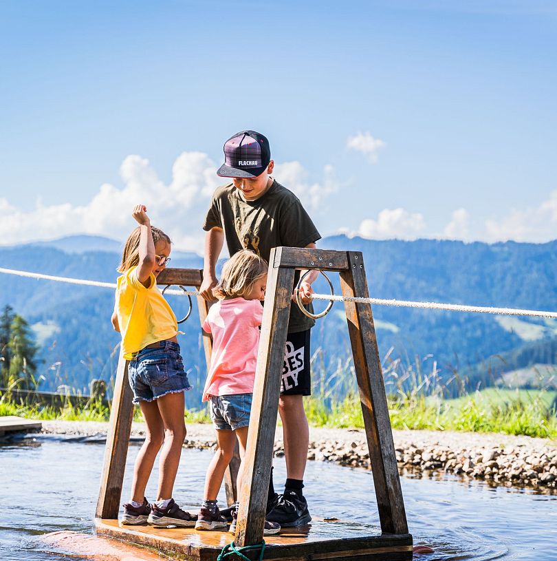Kinder überqueren auf dem Floß den kleinen See - eine der beliebtesten Spielstationen auf Flori's Erlebnispfad in Flachau.