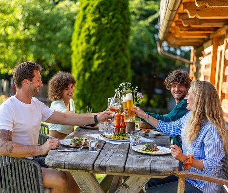 Freunde sitzen an einem Tisch im Garten einer Almhütte in Flachau und stoßen mit Getränken an.
