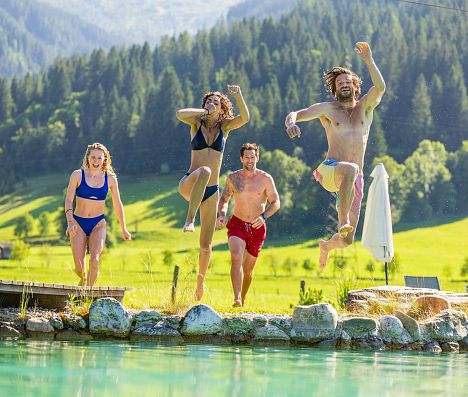 Vier Erwachsene springen lachend von einem Steg in einen Naturbadesee in Flachau, umgeben von grünen Wiesen und Wald.