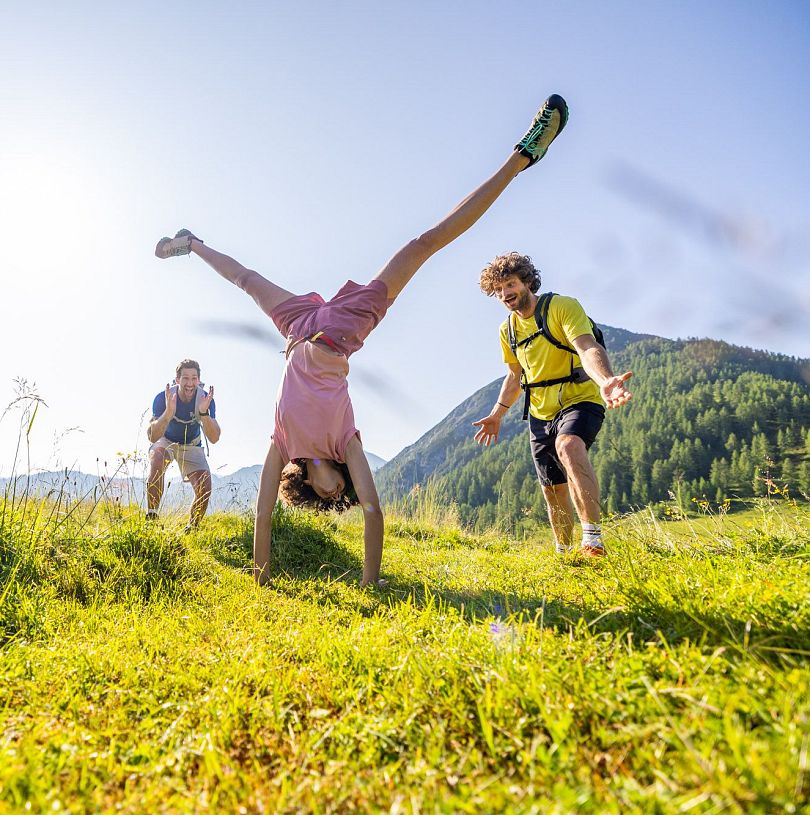 Wandergruppe lacht und jubelt, während eine Person einen Handstand auf einer Almwiese in Flachau macht.