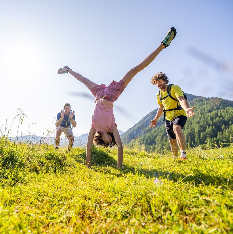 Wandergruppe lacht und jubelt, während eine Person einen Handstand auf einer Almwiese in Flachau macht.