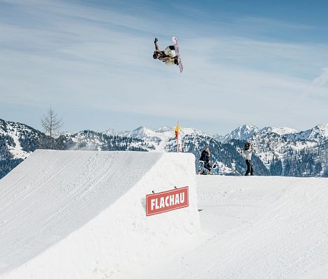 Snowboarder in der Luft nach Absprung auf einer Schanze im Absolut Park in Flachau. Flachau Logo auf Schanze erkennbar.
