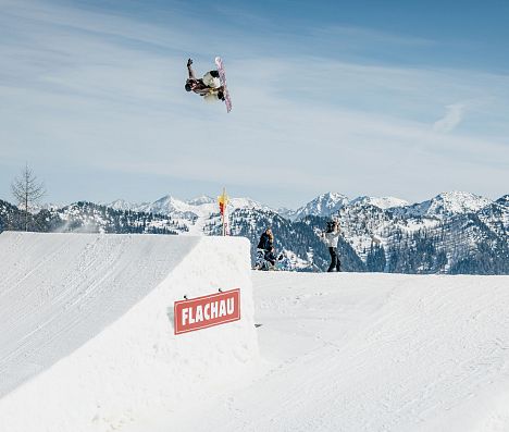Snowboarder in der Luft nach Absprung auf einer Schanze im Absolut Park in Flachau. Flachau Logo auf Schanze erkennbar.