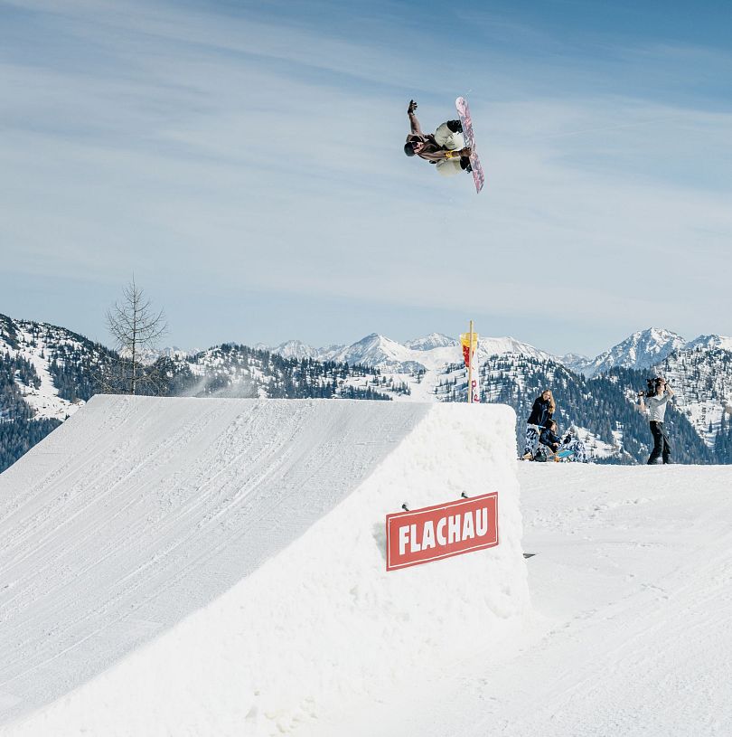 Snowboarder in der Luft nach Absprung auf einer Schanze im Absolut Park in Flachau. Flachau Logo auf Schanze erkennbar.