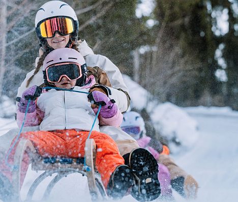 Mutter sitzt mit ihrer Tochter auf einem Schlitten und sie fahren schnell den Berg in Flachau hinunter. Beide tragen einen Helm.