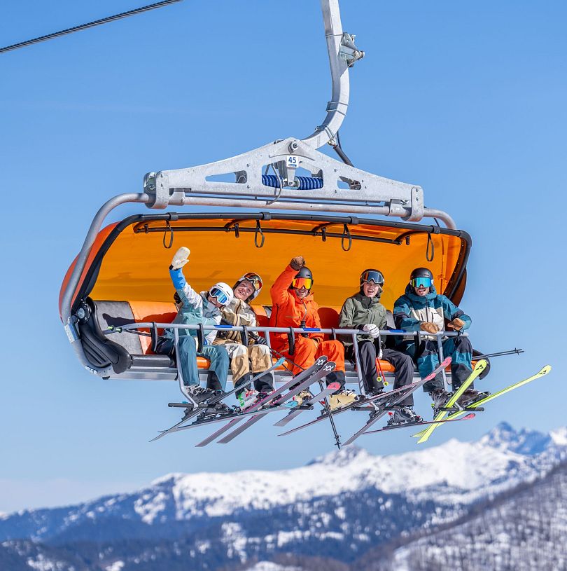 Gruppe von Skifahrern fährt im Sessellift mit orangener Wetterschutzhaube, strahlende Gesichter bei blauem Himmel und verschneiten Bergen. Skigenuss in Flachau.