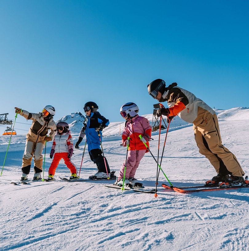Familie steht in Skiausrüstung etwas unterhalb der Bergstation Starjet 3 und bereitet sich auf die Abfahrt vor.