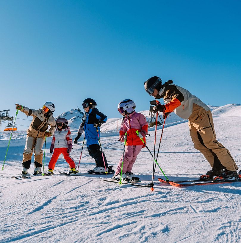 Familie steht in Skiausrüstung etwas unterhalb der Bergstation Starjet 3 und bereitet sich auf die Abfahrt vor.