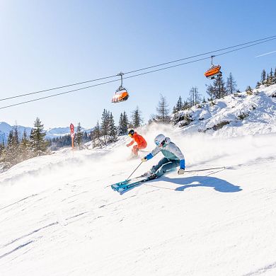 Zwei Skifahrer bei der Abfahrt vor einem orangen Sessellift. Beste Pistenverhältnisse und blauer Himmel.