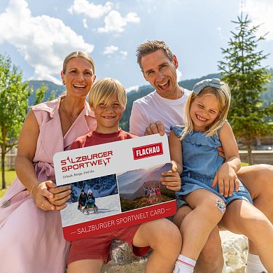 Eine Familie mit zwei Kindern hält eine grosse Version der Flachau Sommer Card in der Hand und lacht in die Kamera.