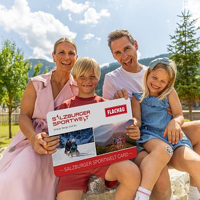 Eine Familie mit zwei Kindern hält eine grosse Version der Flachau Sommer Card in der Hand und lacht in die Kamera.