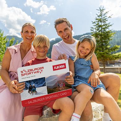 Eine Familie mit zwei Kindern hält eine grosse Version der Flachau Sommer Card in der Hand und lacht in die Kamera.