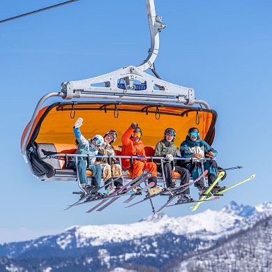 Gruppe von Skifahrern fährt im Sessellift mit orangener Wetterschutzhaube, strahlende Gesichter bei blauem Himmel und verschneiten Bergen. Skigenuss in Flachau.