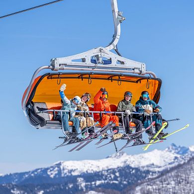 Gruppe von Skifahrern fährt im Sessellift mit orangener Wetterschutzhaube, strahlende Gesichter bei blauem Himmel und verschneiten Bergen. Skigenuss in Flachau.